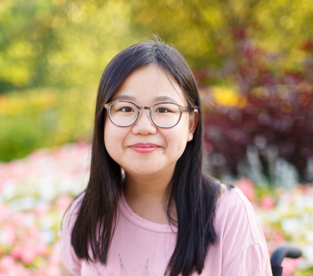 A picture of Amy, a disabled Chinese-Canadian woman sitting in a manual wheelchair. She has long, straight, black hair. She is wearing a pink t-shirt and silver glasses. The background consists of blurred trees, and pink and white flowers. She is smiling at the camera.
