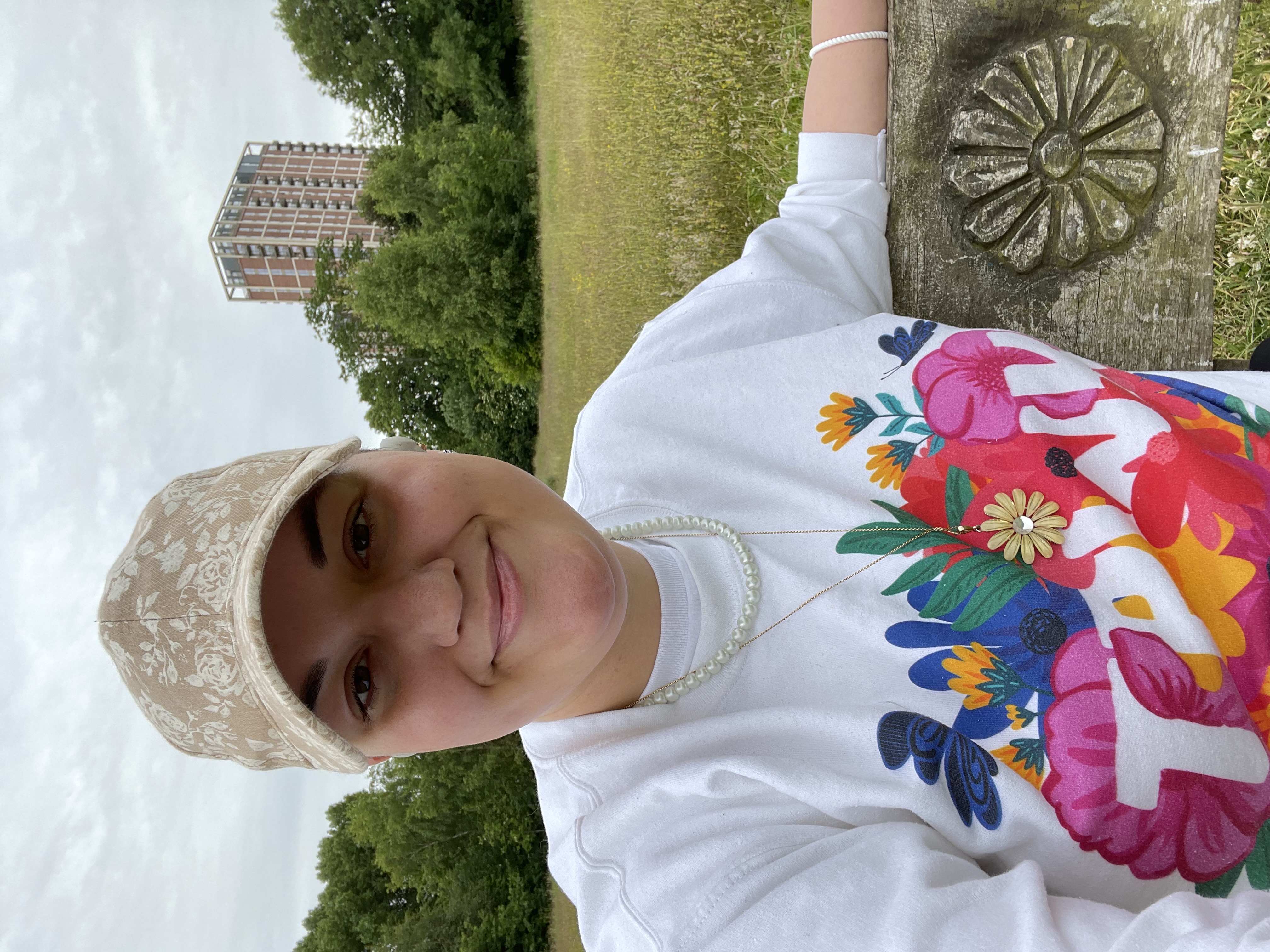Photo of Andy Zubac, a white non-binary person, smiling into the camera while sitting on a bench in the middle of a grassy field. They are wearing a hat, sweater, and jewellery all following a flower motif and the bench is also carved with a matching flower design.